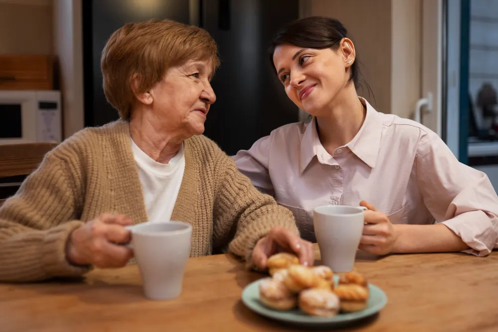 A companion carer and an elderly woman smile at each other over cups of tea and pastries at a kitchen table during a home visit