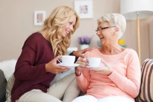 A visiting carer and an older woman share a cup of tea and a laugh on the sofa during an hourly home care visit