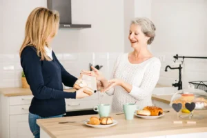 A live-in carer serves breakfast to a smiling older woman in a bright home kitchen in London