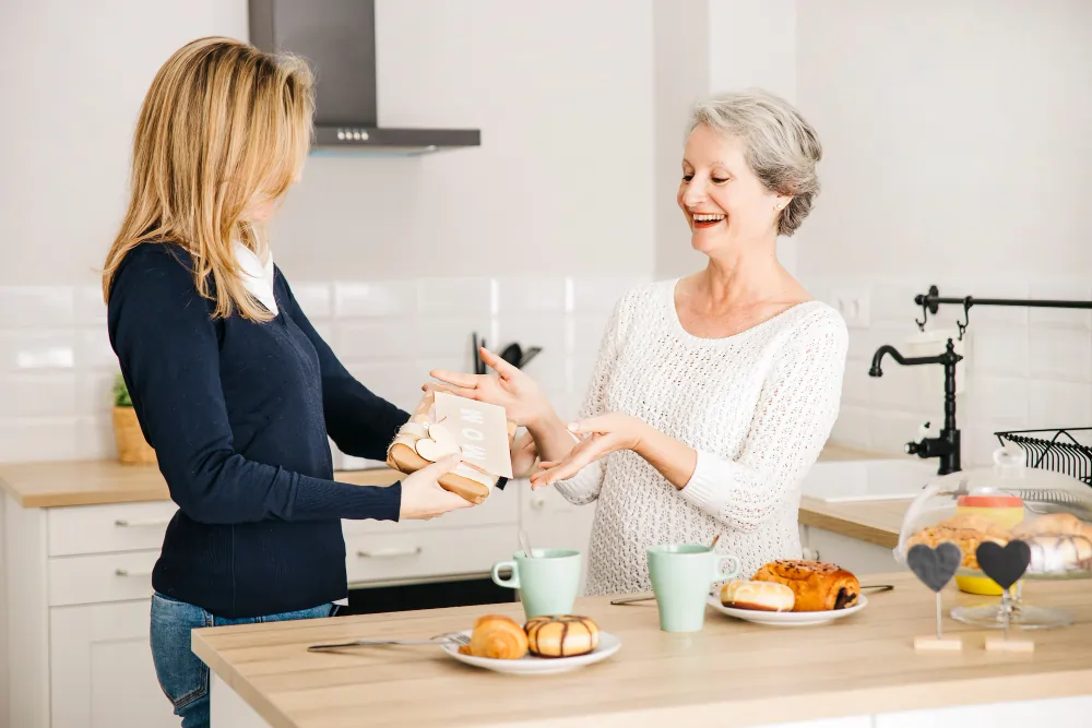 A live-in carer serves breakfast to a smiling older woman in a bright home kitchen in London